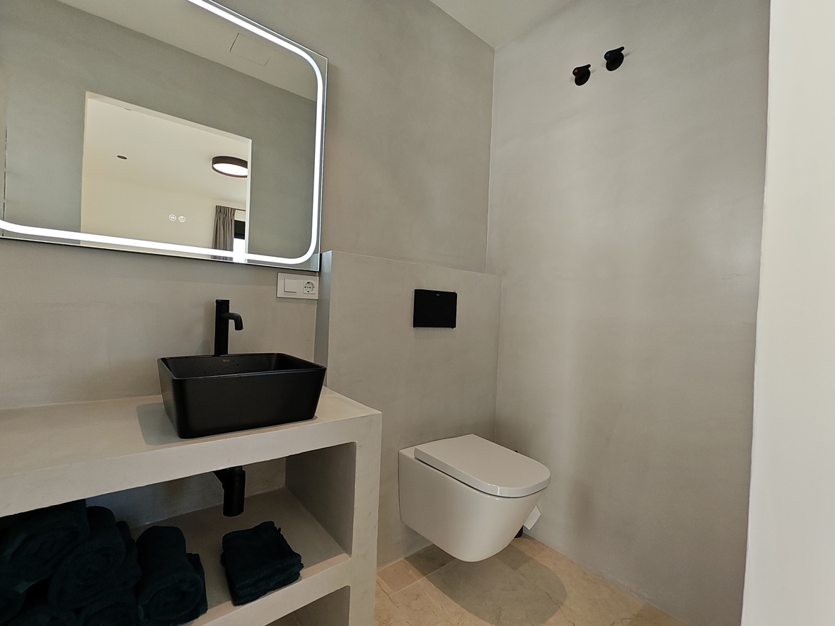 Salle d'eau moderne avec vasque noire, miroir rétroéclairé à bords arrondis et WC suspendu sur un revêtement mural en microciment.