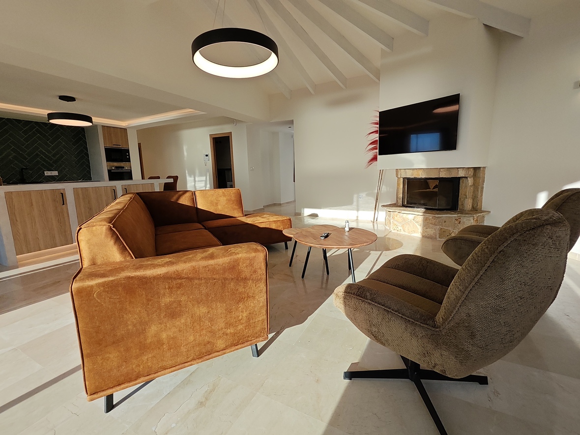 Salon contemporain avec canapé en velours ocre, cheminée en pierre et plafond voûté. La cuisine ouverte à l'arrière-plan présente une crédence verte et des finitions en bois clair.