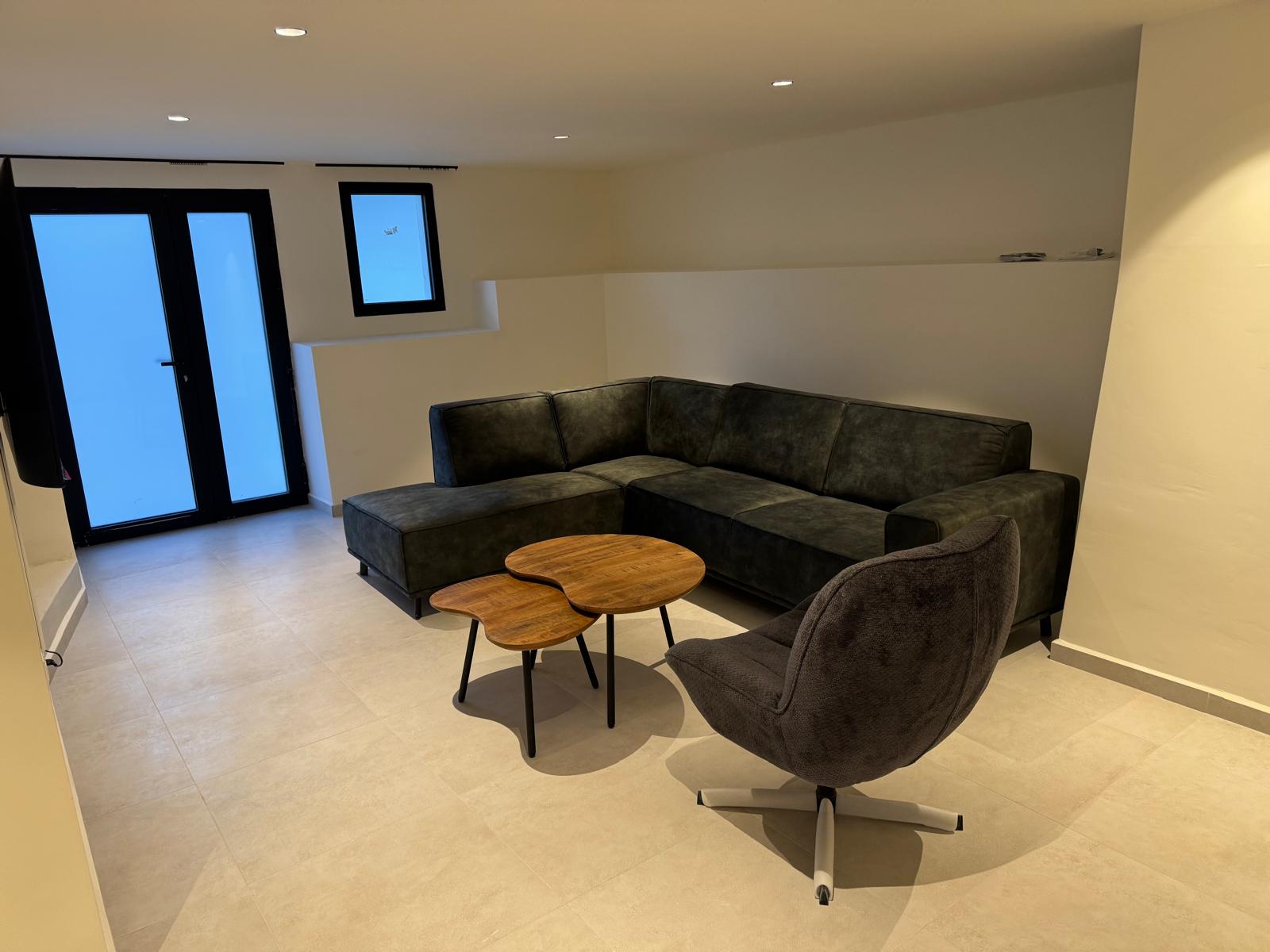 Salon moderne avec canapé d'angle gris foncé, tables basses en bois de forme organique et fauteuil pivotant sur un sol en carrelage clair et spots encastrés.