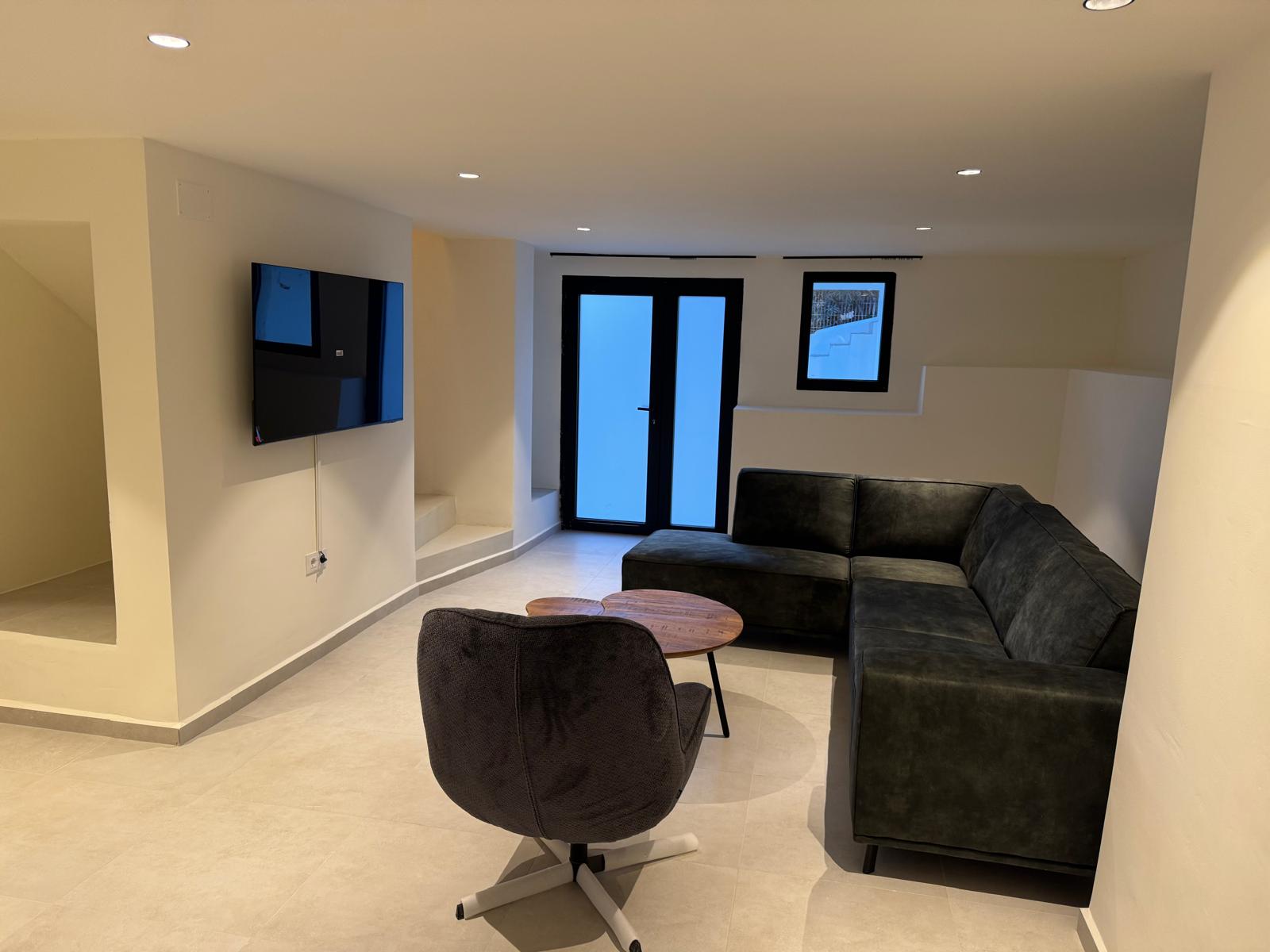 Salon moderne avec canapé d'angle sombre, fauteuil pivotant et TV murale. L'éclairage encastré et le sol neutre soulignent l'esthétique épurée de cet espace.