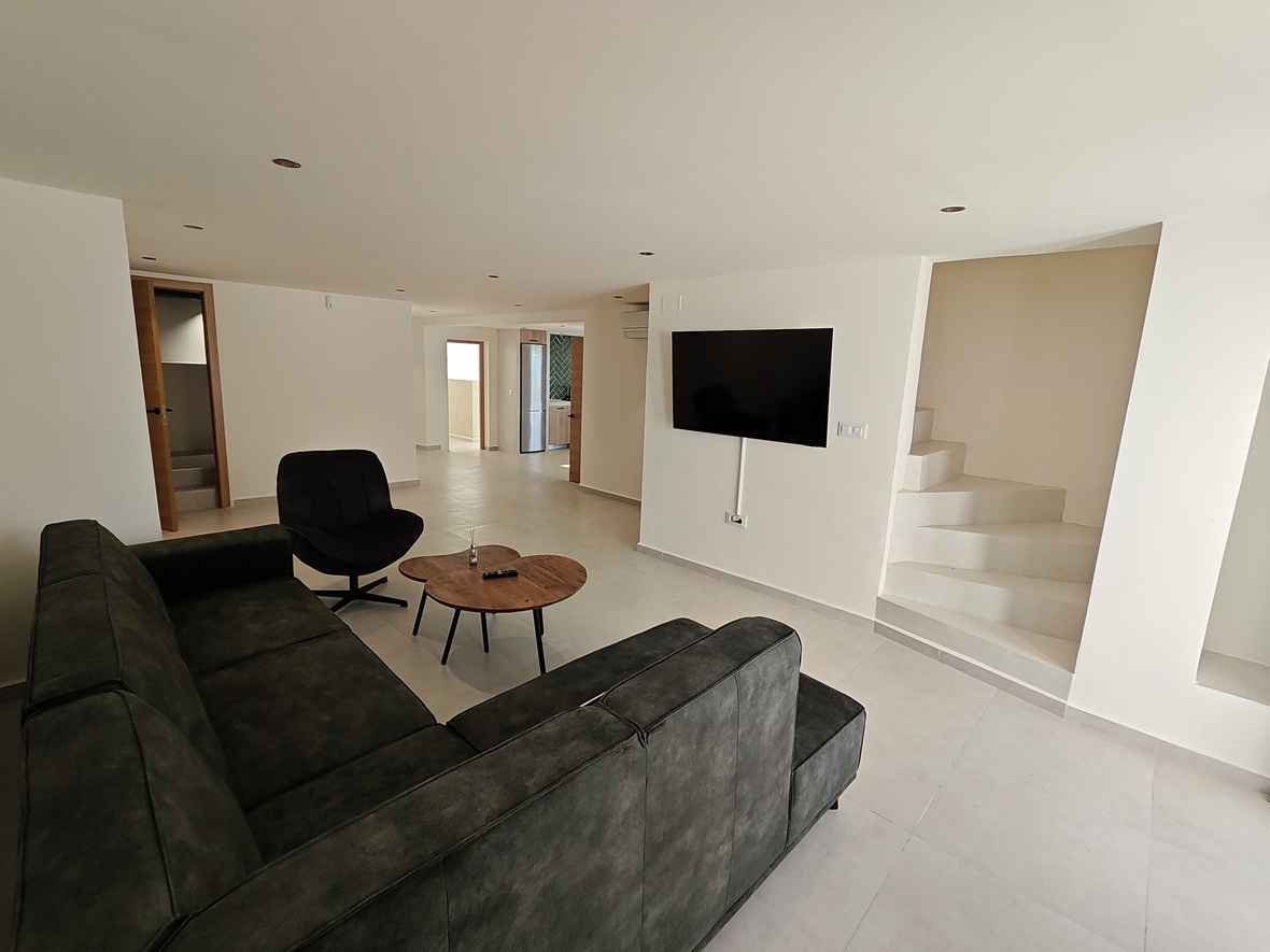 Salon moderne avec canapé d'angle sombre, table basse en bois et TV murale, jouxtant un escalier blanc minimaliste et des spots encastrés.
