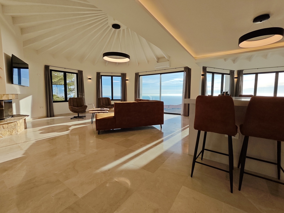 Salon ouvert avec plafond à poutres radiales, sol en marbre poli et vues panoramiques sur la mer. Comprend une cheminée en pierre et un éclairage circulaire moderne.