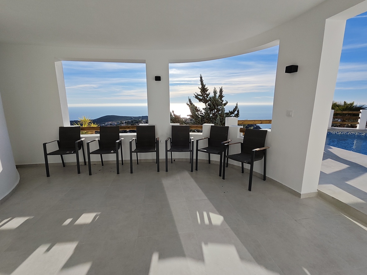 Terrasse couverte avec fauteuils noirs modernes, carrelage gris clair et larges ouvertures offrant une vue panoramique sur la mer et l'espace piscine attenant.