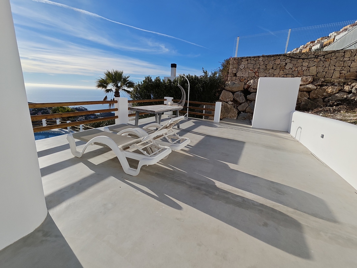 Terrasse ensoleillée avec chaises longues blanches, table en bois et fauteuil suspendu. Vue panoramique sur la mer, murs en pierre et sol en béton lissé.