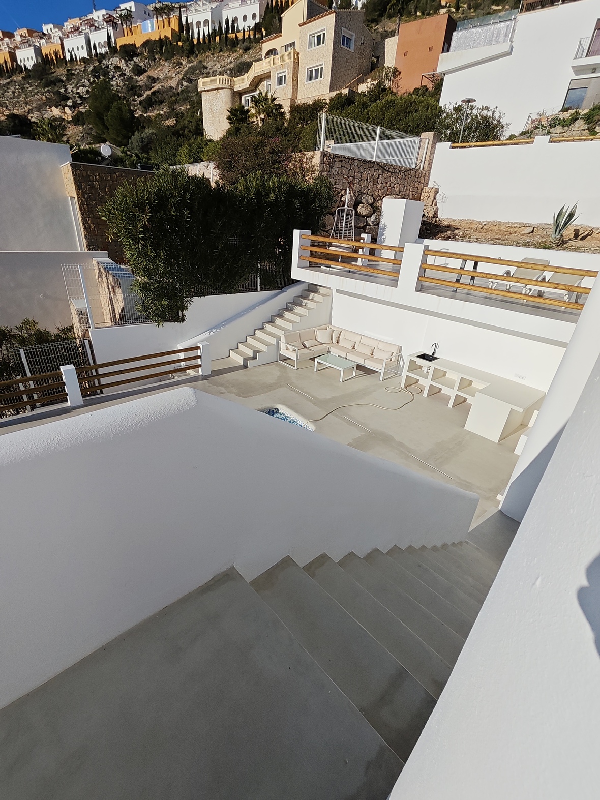 Terrasse extérieure à plusieurs niveaux avec murs blancs, garde-corps en bois, coin salon avec canapé d'angle et cuisine d'été intégrée sur fond de colline méditerranéenne.
