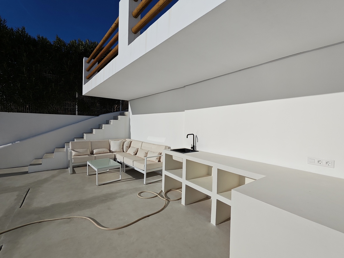 Terrasse extérieure avec cuisine d'été maçonnée en blanc, évier noir et coin salon composé d'un canapé d'angle et d'une table basse.
