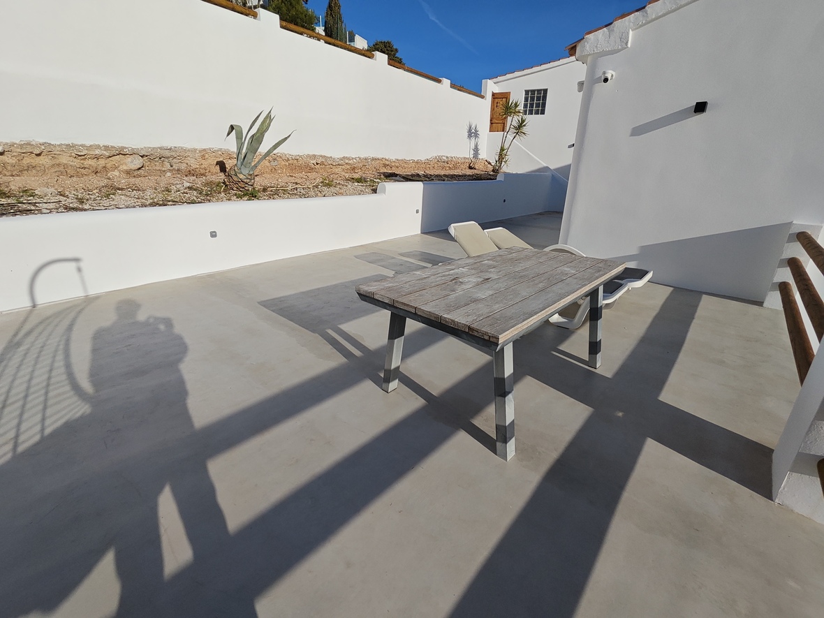 Terrasse extérieure en microciment avec table en bois patiné, entourée de murs blancs et d'une végétation méditerranéenne.