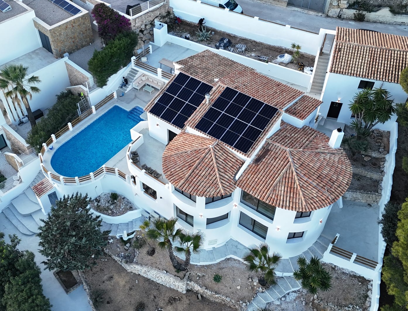 Vue aérienne d'une villa méditerranéenne aux toits en terre cuite équipés de panneaux solaires, avec piscine ovale et terrasses en pierre sur plusieurs niveaux.