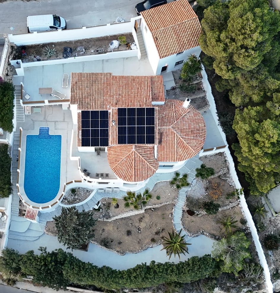 Vue aérienne d'une villa méditerranéenne avec panneaux solaires sur toit en terre cuite, piscine incurvée et jardins en terrasses avec plantes locales.
