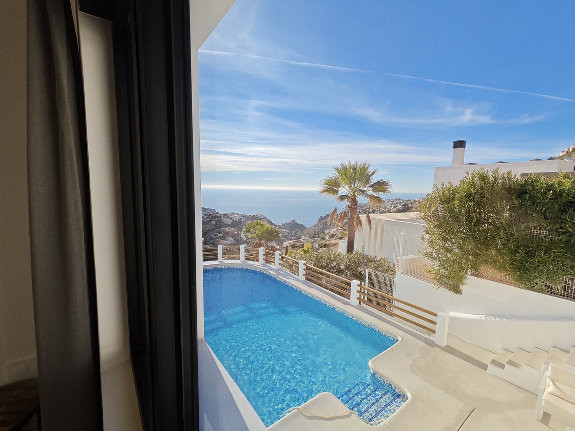 Vue de l'intérieur sur une piscine en mosaïque bleue et une terrasse blanche surplombant la côte méditerranéenne et des palmiers sous un ciel dégagé.