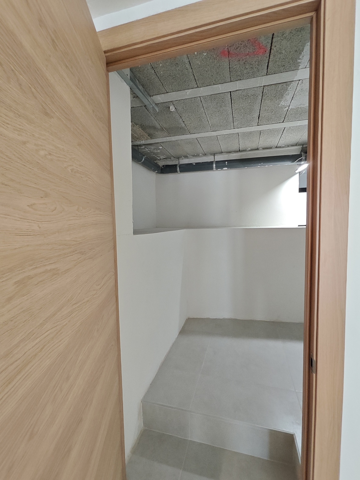 Vue d'un local technique ou de rangement avec porte en bois clair, carrelage gris au sol et plafond brut avec canalisations apparentes.