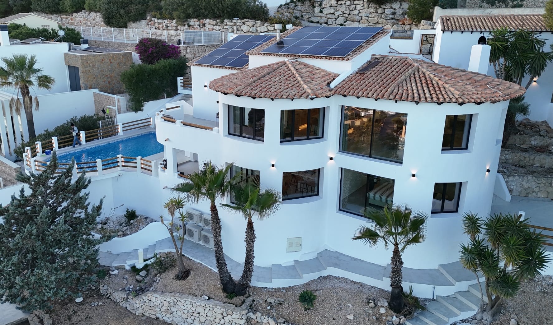 Vue extérieure d'une villa blanche à plusieurs niveaux avec architecture courbe, toits en tuiles, panneaux solaires et piscine privée bordée de palmiers.