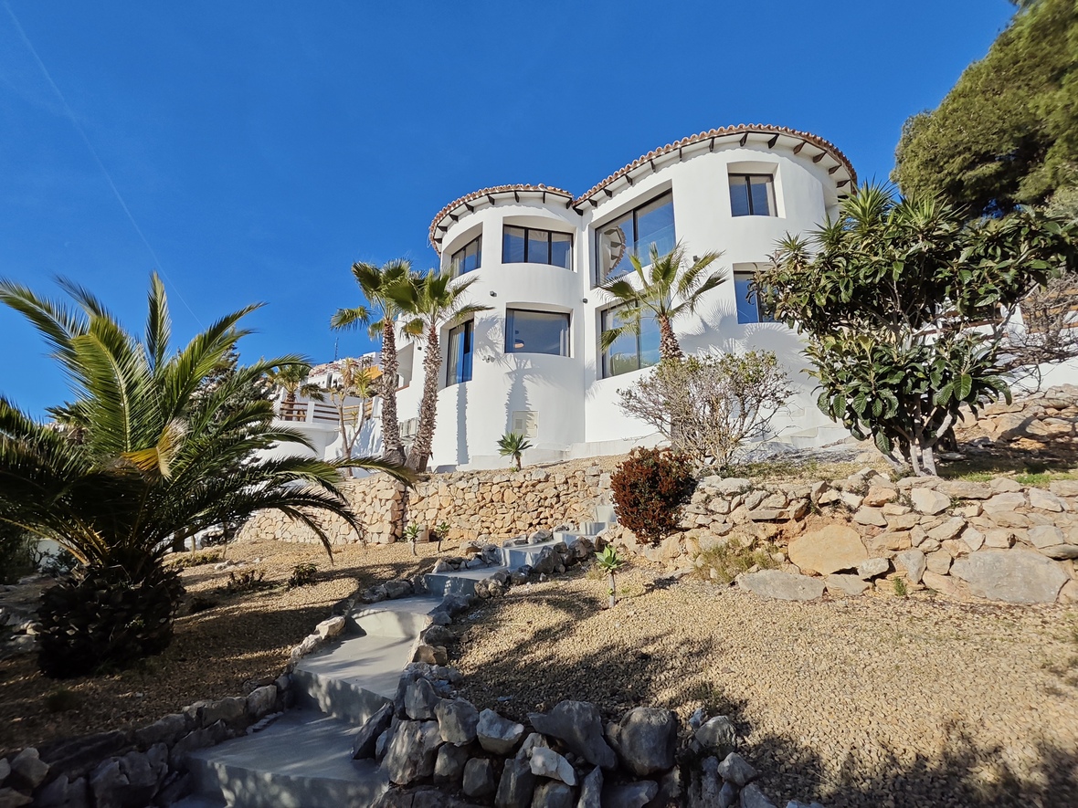 Vue extérieure d'une villa blanche aux lignes courbes et larges baies vitrées. Le jardin en pente dispose de murs en pierre, de palmiers et d'un escalier d'accès en béton.