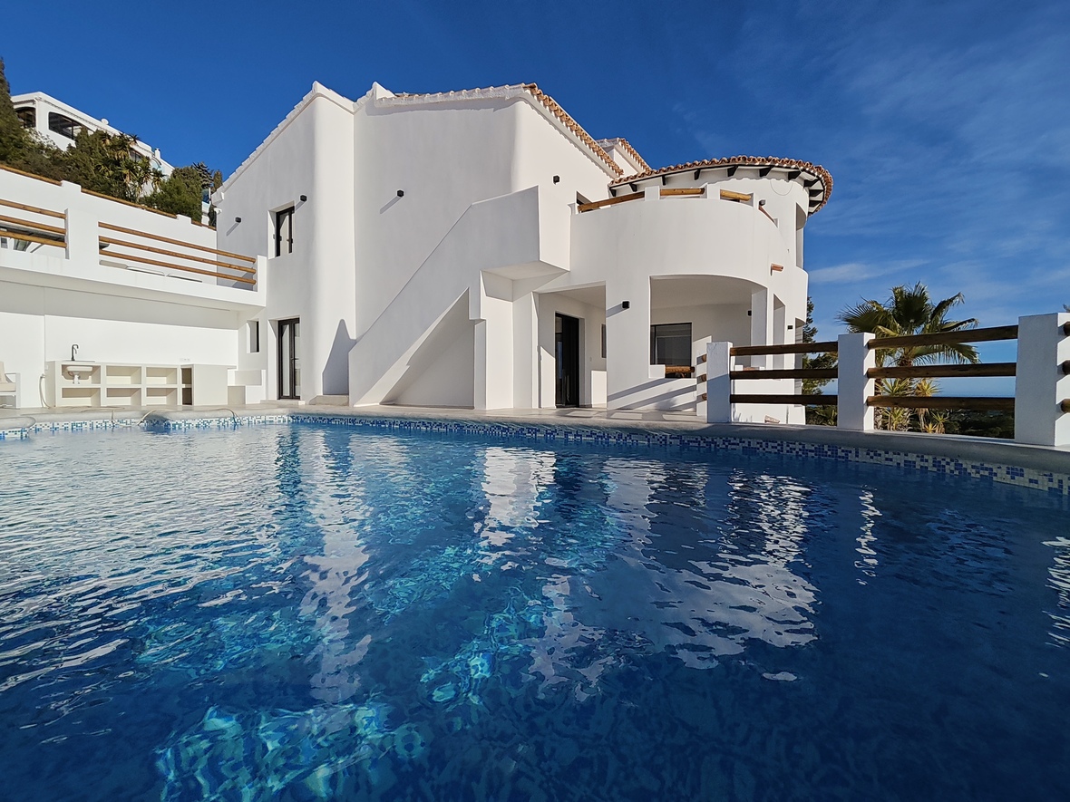 Vue extérieure d'une villa blanche aux terrasses courbes, garde-corps en bois et piscine en mosaïque bleue reflétant la structure méditerranéenne.