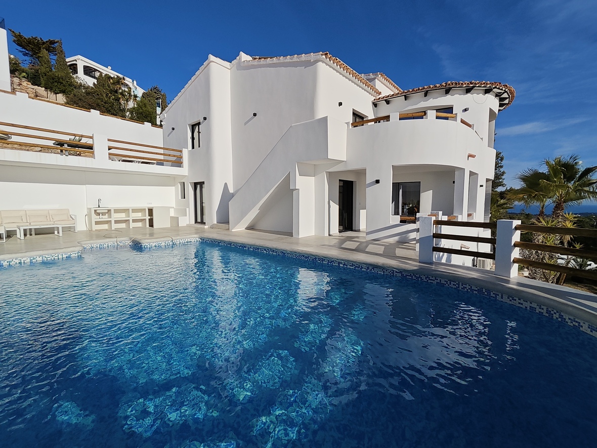 Vue extérieure d'une villa blanche avec piscine incurvée en mosaïque bleue, terrasses à plusieurs niveaux et garde-corps en bois.