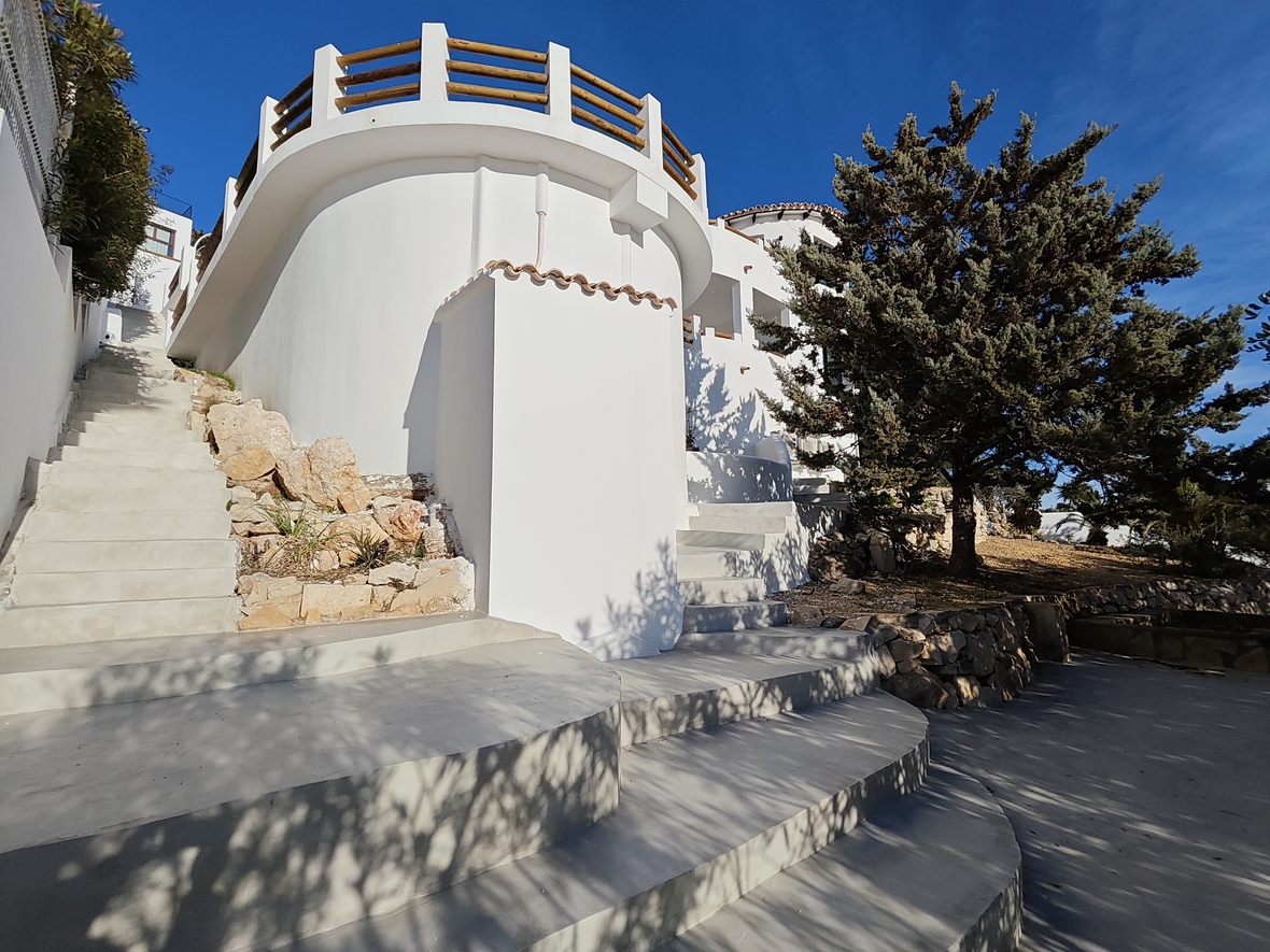 Vue extérieure d'une villa méditerranéenne aux murs blancs arrondis, escaliers en béton et terrasse circulaire avec garde-corps en bois.
