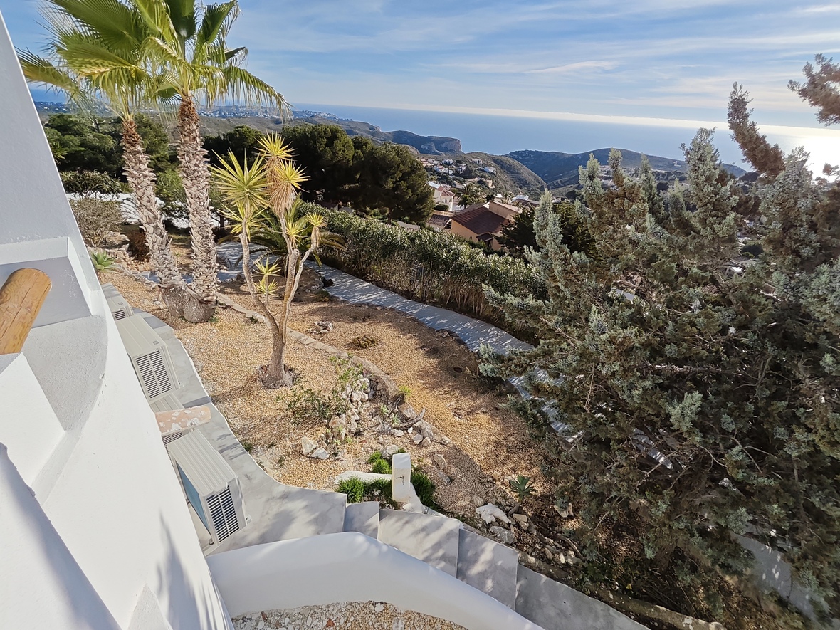 Vue surélevée d'un jardin paysager avec palmiers et allées de gravier, offrant un panorama sur la vallée côtière et la mer Méditerranée.
