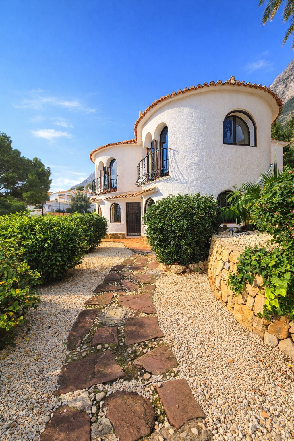 Façade de villa méditerranéenne aux murs blancs arrondis, fenêtres cintrées et balcons en fer forgé. Allée en pierre naturelle entourée de gravier et végétation.