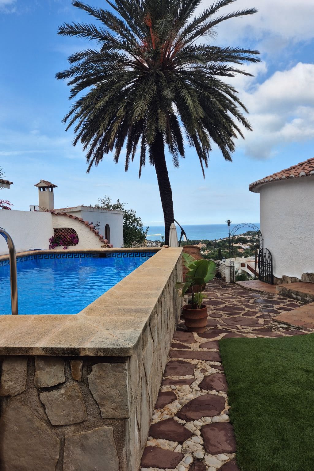 Piscine surélevée avec revêtement en pierre, terrasse en dalles irrégulières, grand palmier et vue dégagée sur la mer Méditerranée.