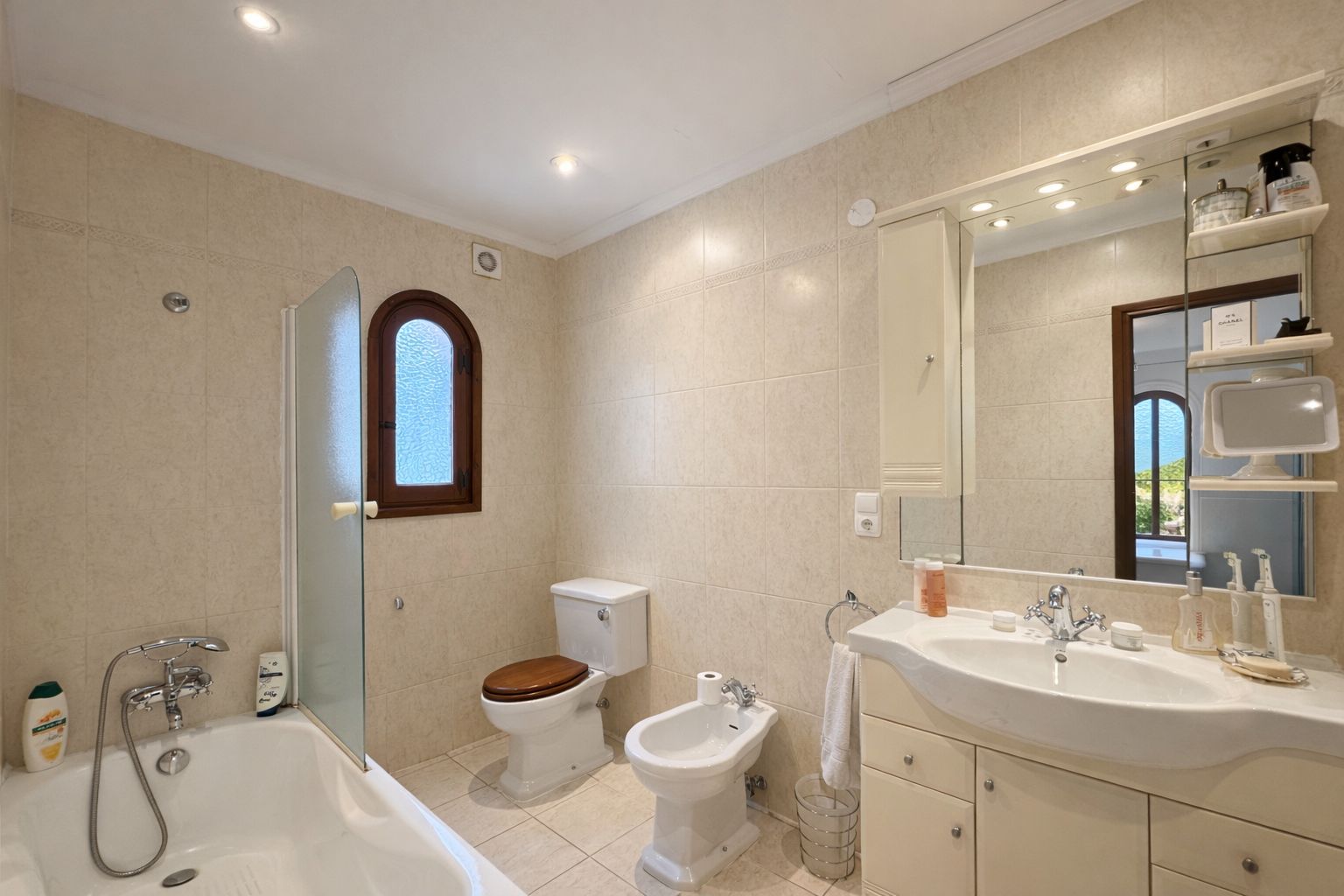 Salle de bain avec baignoire, paroi en verre, bidet et toilettes à abattant en bois. Comprend un meuble vasque, un grand miroir éclairé et une fenêtre cintrée.