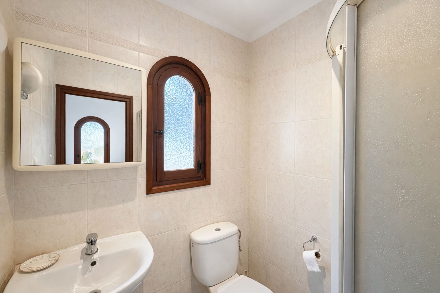 Salle de bain avec carrelage mural beige, fenêtre cintrée en bois à verre texturé et lavabo en céramique blanche.