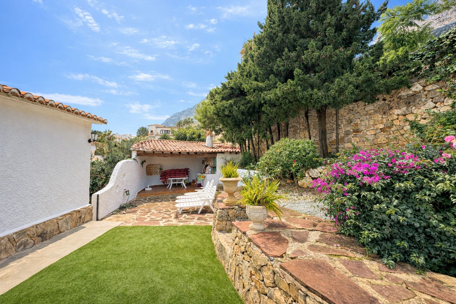 Terrasse extérieure avec pavage en pierre naturelle, gazon artificiel et porche couvert. Des murs en pierre sèche et une végétation méditerranéenne entourent l'espace.