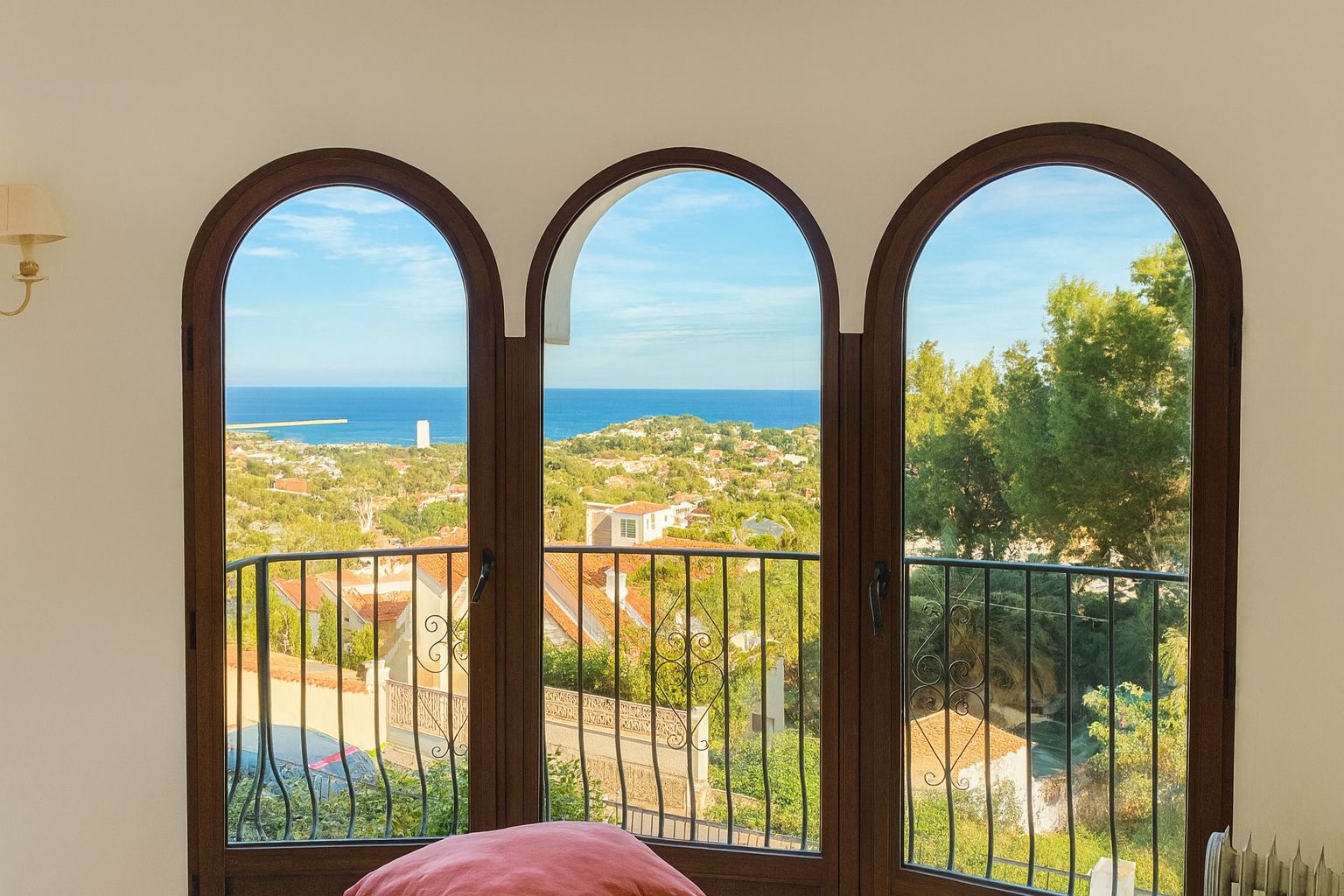 Trois fenêtres cintrées avec cadres en bois sombre offrent une vue panoramique sur la mer Méditerranée et le littoral, avec un garde-corps extérieur en fer forgé.