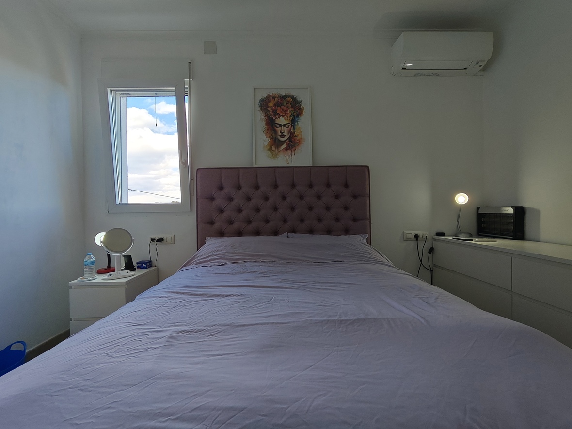 Chambre à coucher avec tête de lit capitonnée mauve, mobilier blanc épuré et climatisation murale. Une fenêtre apporte une luminosité naturelle à la pièce.