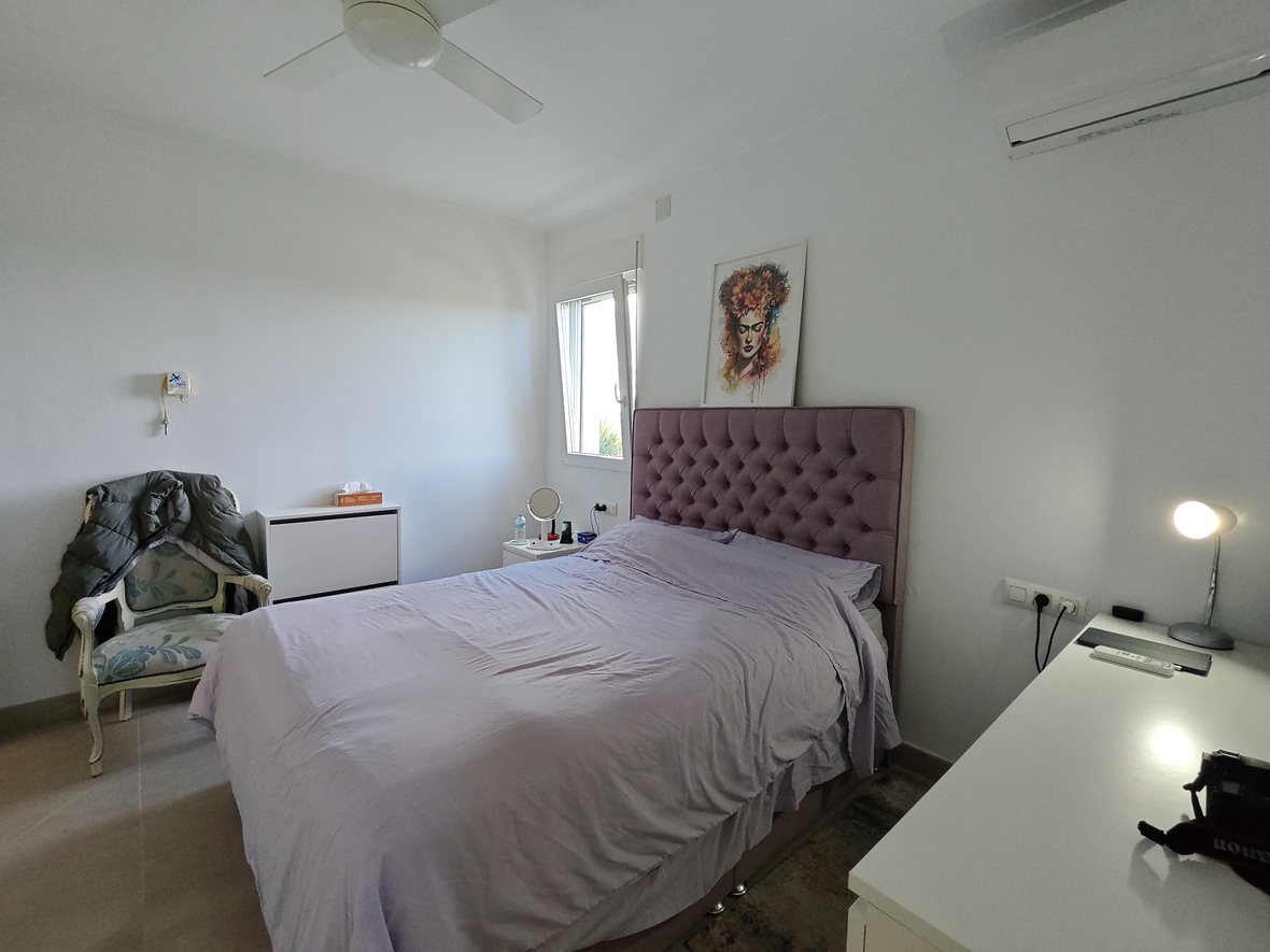 Chambre à coucher avec tête de lit capitonnée mauve, murs blancs, ventilateur de plafond et un coin bureau pratique avec lumière naturelle.