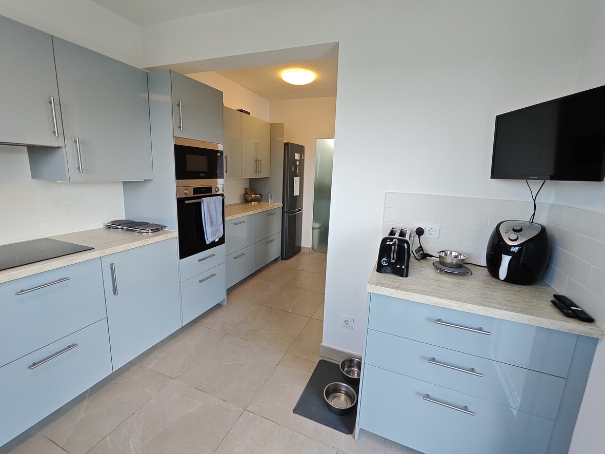 Cuisine moderne avec meubles bleu clair, appareils intégrés et plans de travail effet pierre. Comprend une télévision murale et un carrelage au sol gris.