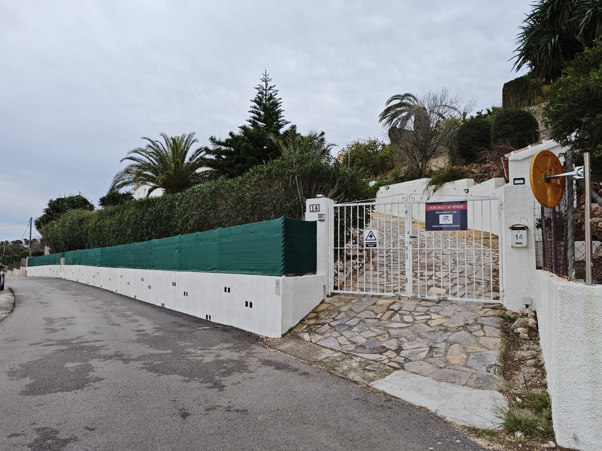 Entrée principale avec portail métallique blanc, allée pavée en pierre naturelle et mur d'enceinte bordé de palmiers et de pins.
