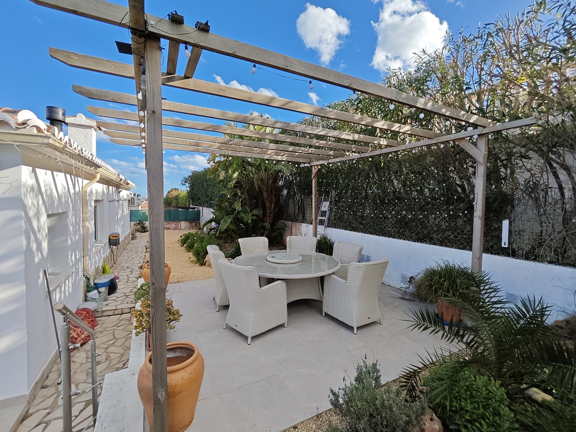 Espace repas extérieur avec table ronde en verre et six chaises en osier blanc sous une pergola en bois agrémentée de guirlandes lumineuses et de plantes.