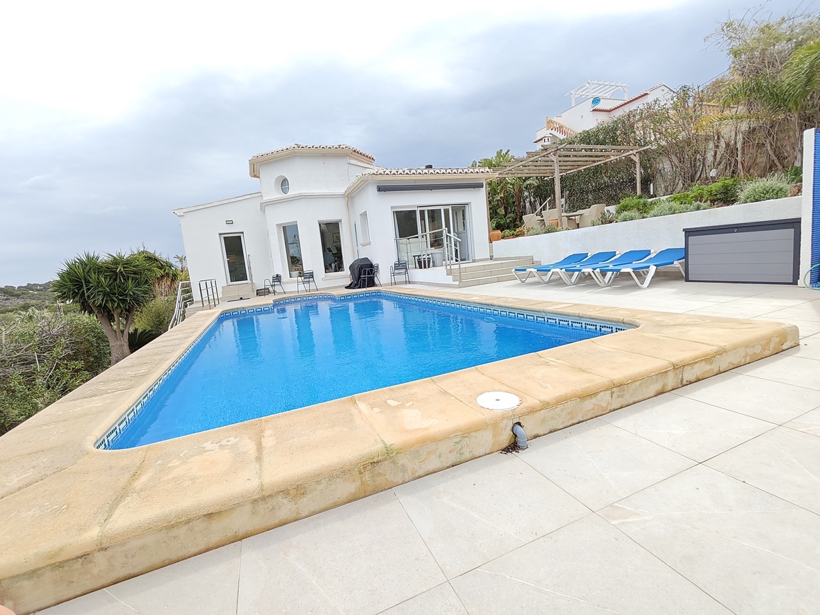 Piscine privée rectangulaire avec carrelage bleu et bordure en pierre, entourée d'une terrasse dallée, de chaises longues et d'une villa blanche méditerranéenne.