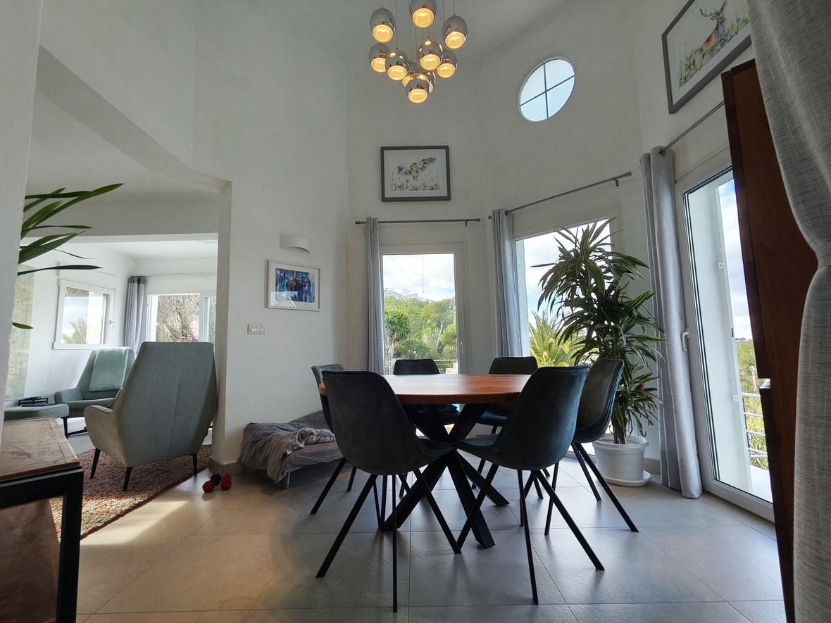 Salle à manger moderne avec table ronde en bois, chaises sombres, hauts plafonds, fenêtre circulaire et lustre chromé à plusieurs globes.