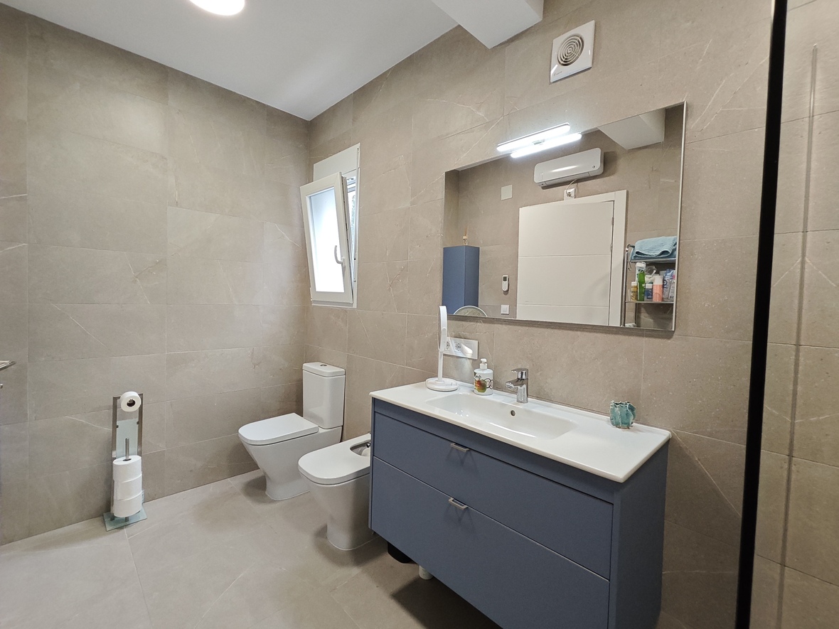Salle de bain moderne avec carrelage en grès cérame beige, meuble vasque suspendu bleu et sanitaires complets. Un grand miroir avec éclairage LED complète l'espace.