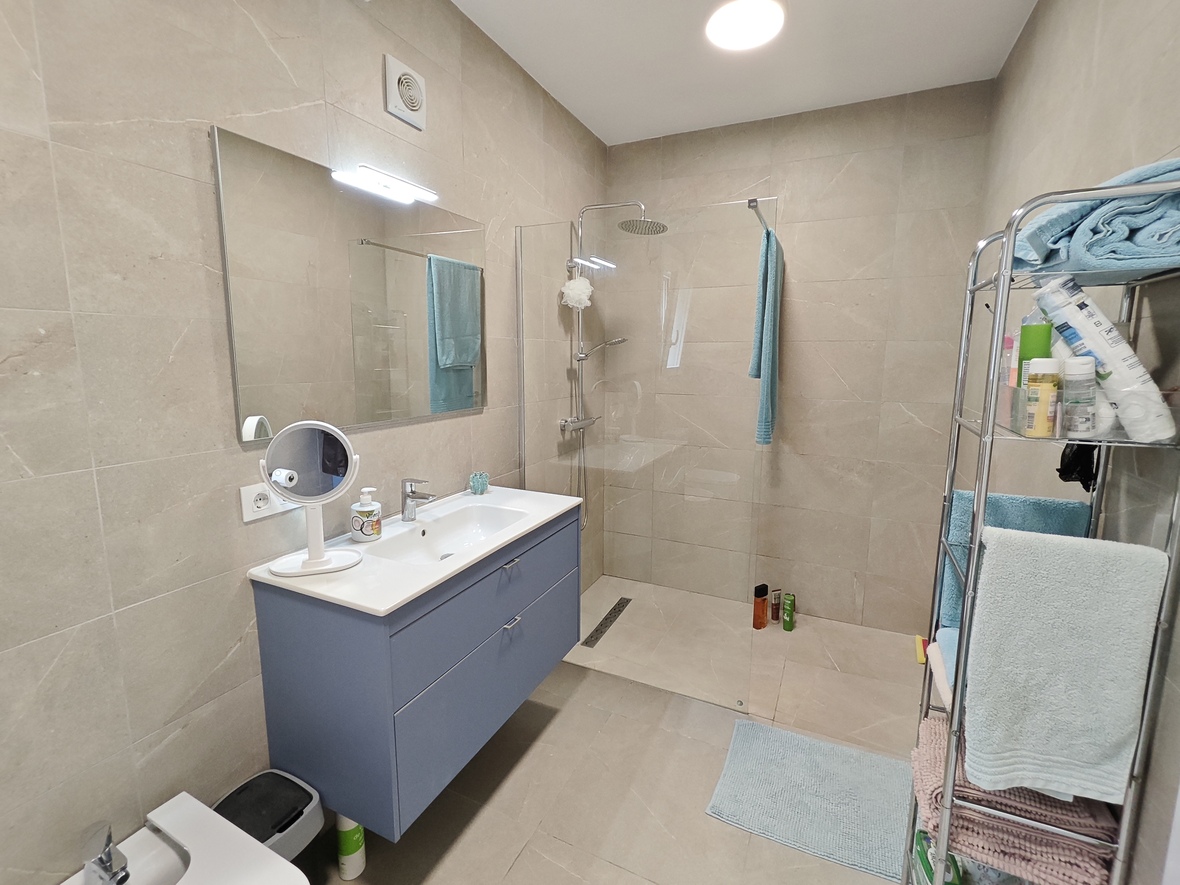 Salle de bain moderne avec meuble vasque bleu suspendu, douche à l'italienne avec paroi vitrée et carrelage mural beige effet pierre.