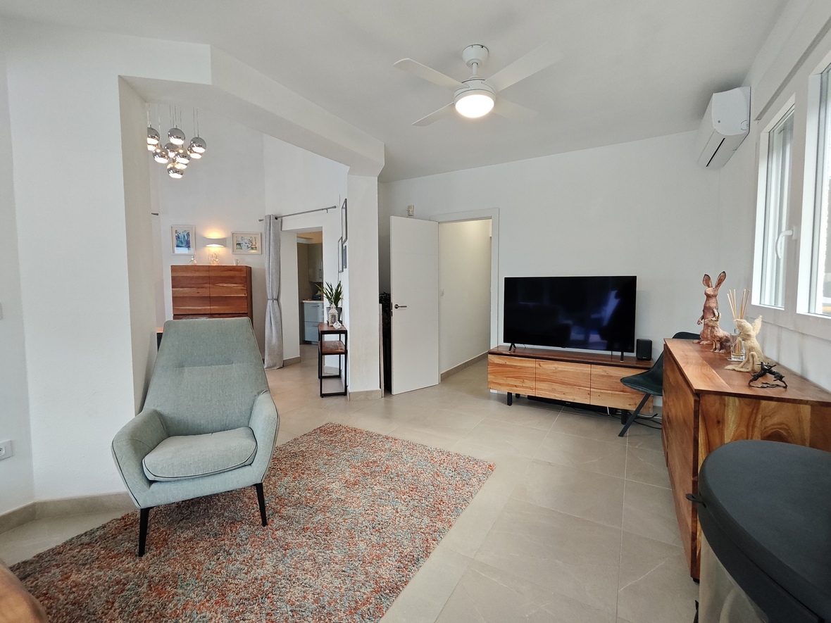 Salon lumineux avec fauteuil moderne, meubles en bois naturel et télévision à écran plat. Équipé d'un ventilateur de plafond, de la climatisation et d'un sol en carrelage clair.