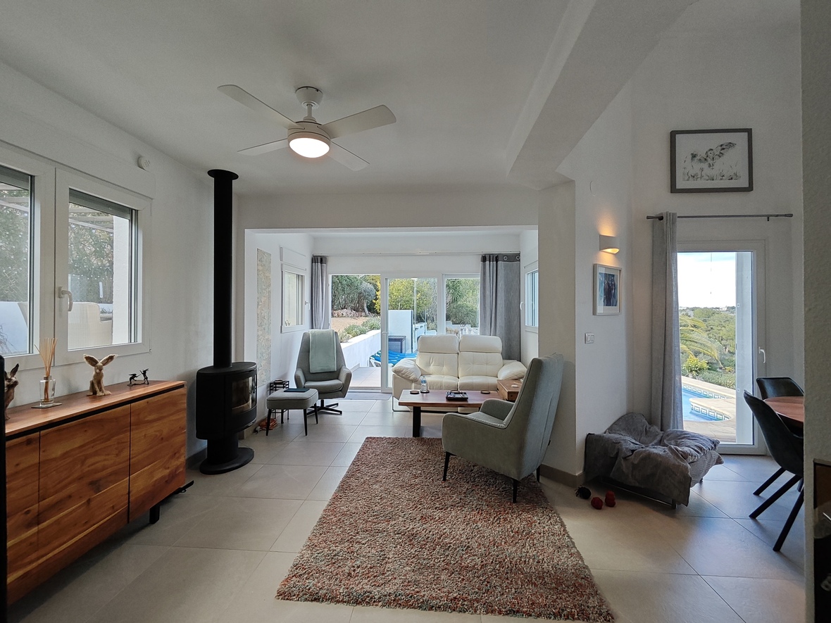 Salon lumineux avec poêle à bois noir, buffet en bois et ventilateur de plafond. De grandes baies vitrées s'ouvrent sur la terrasse et la piscine.