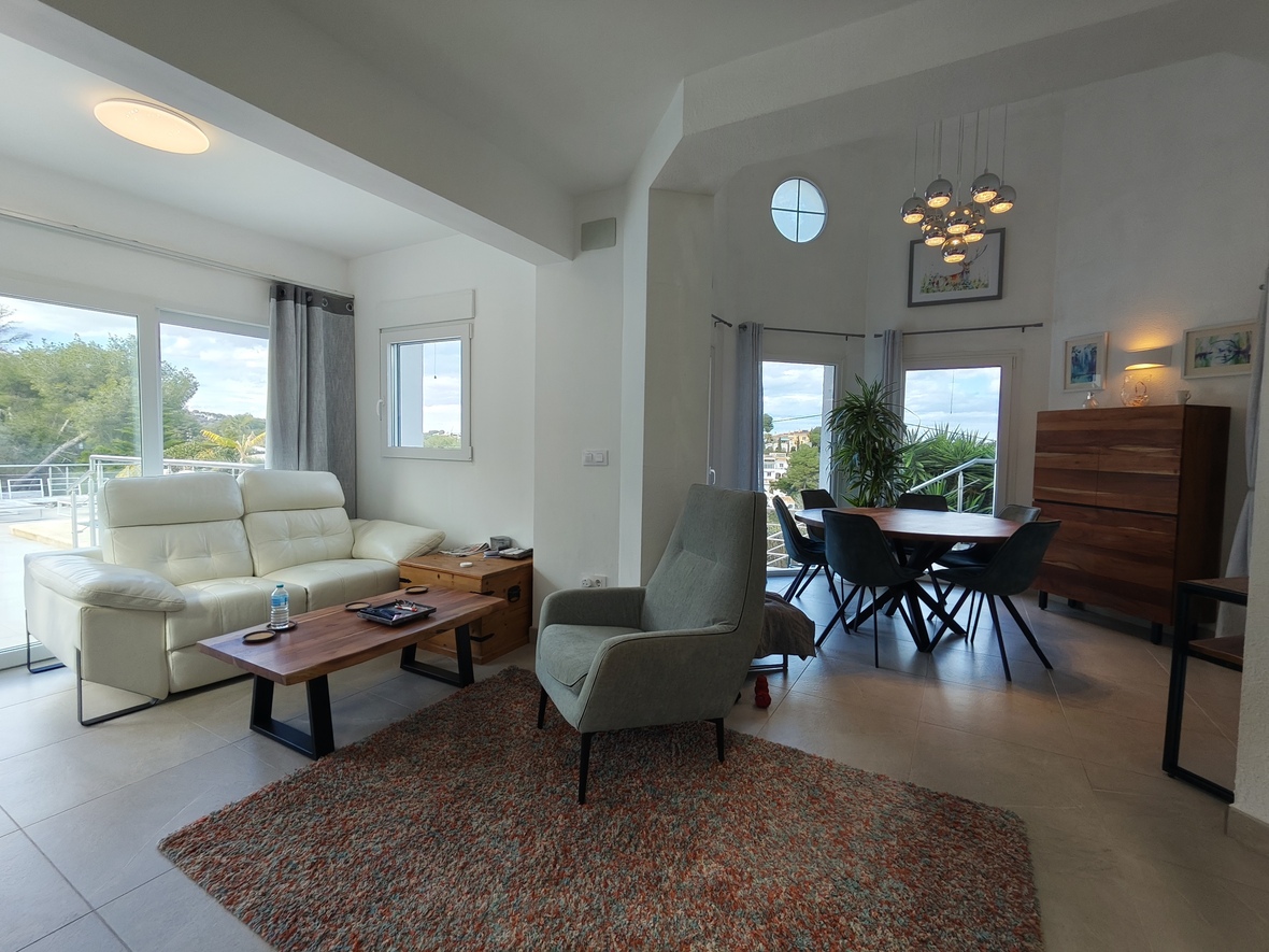 Séjour ouvert avec salon en cuir blanc et coin repas doté d'un luminaire design. Les grandes ouvertures vitrées apportent une luminosité naturelle et un accès à la terrasse.