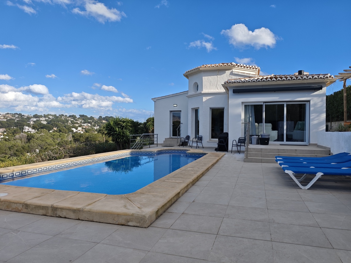 Terrasse extérieure avec piscine rectangulaire, bordure en pierre, bains de soleil bleus et vue sur les collines et la végétation.