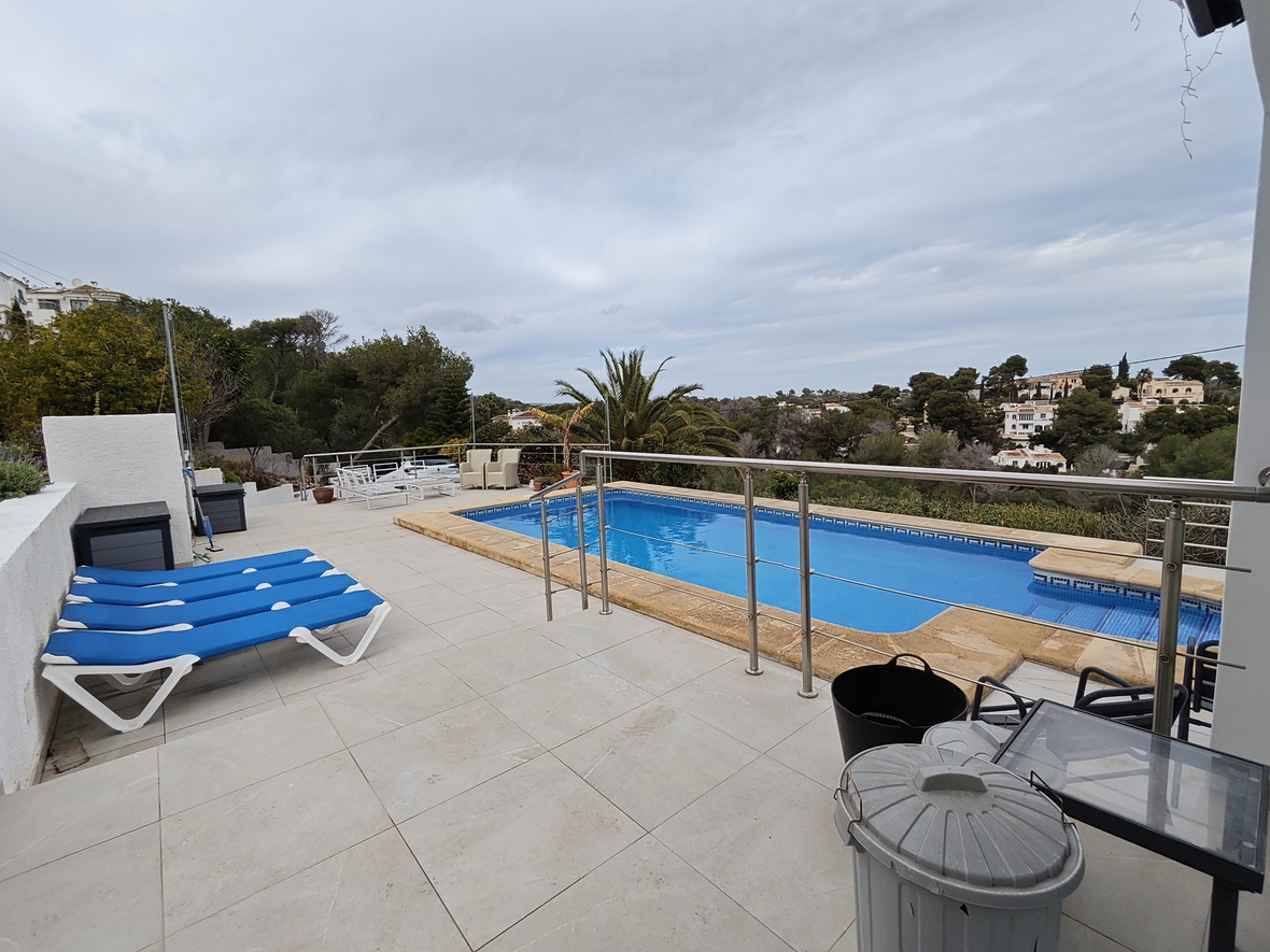 Terrasse surélevée avec piscine rectangulaire, bordures en pierre, chaises longues bleues et garde-corps en inox offrant une vue dégagée sur la vallée boisée.
