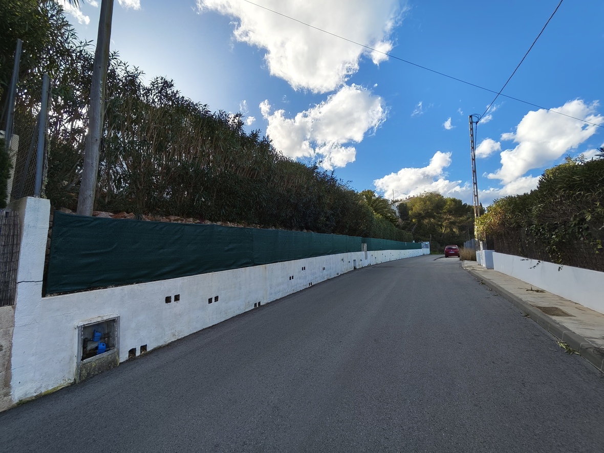 Vue de la rue d'accès résidentielle avec un mur de clôture blanc, un filet de protection vert et des haies denses sous un ciel bleu nuageux.