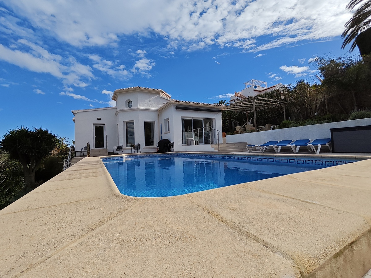 Vue extérieure d'une villa blanche avec piscine privée, terrasse en pierre, chaises longues et architecture méditerranéenne sous un ciel dégagé.