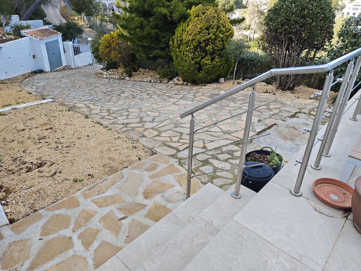 Vue extérieure montrant une allée en pierre naturelle entourée de végétation méditerranéenne et un escalier avec garde-corps en acier inoxydable.