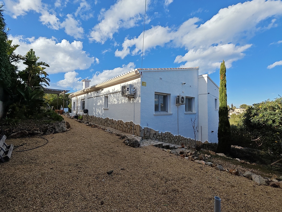 Vue latérale d'une villa blanche avec soubassement en pierre, allée de gravier et climatiseurs muraux, entourée de végétation méditerranéenne et d'un cyprès.