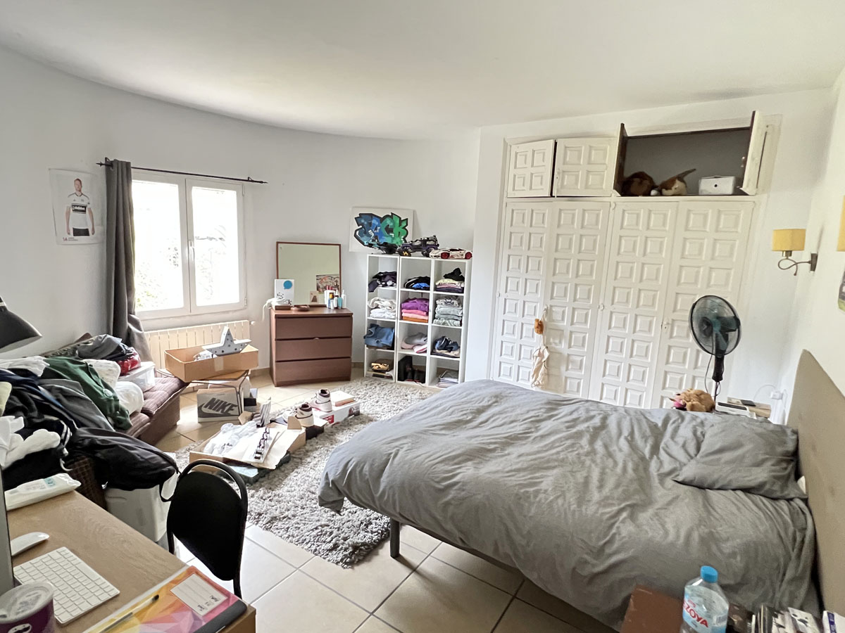 Chambre avec une grande armoire encastrée aux portes blanches travaillées, fenêtre latérale et sol en carrelage. L'espace intègre un coin bureau et du rangement.