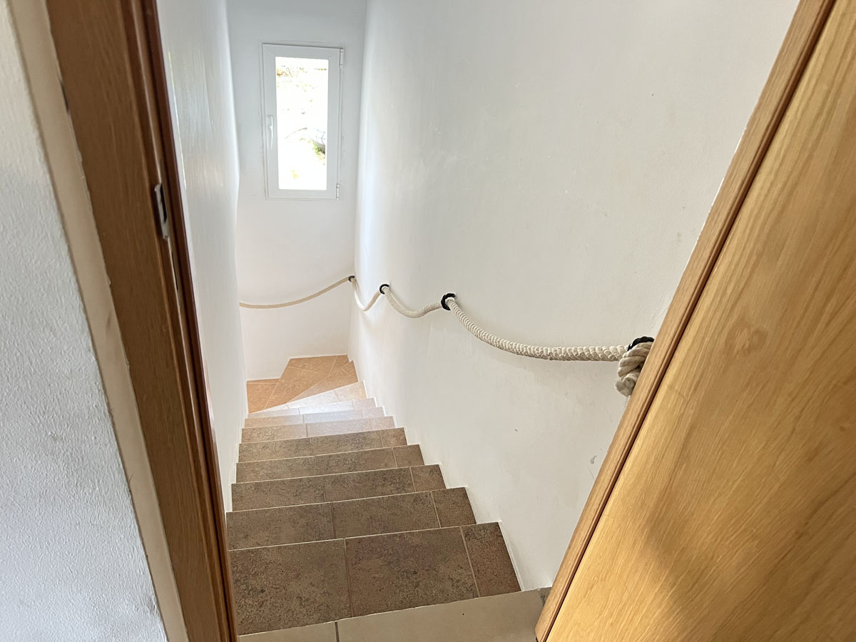 Escalier intérieur avec marches en céramique et une main courante originale en corde, éclairé par une fenêtre verticale sur des murs blancs.