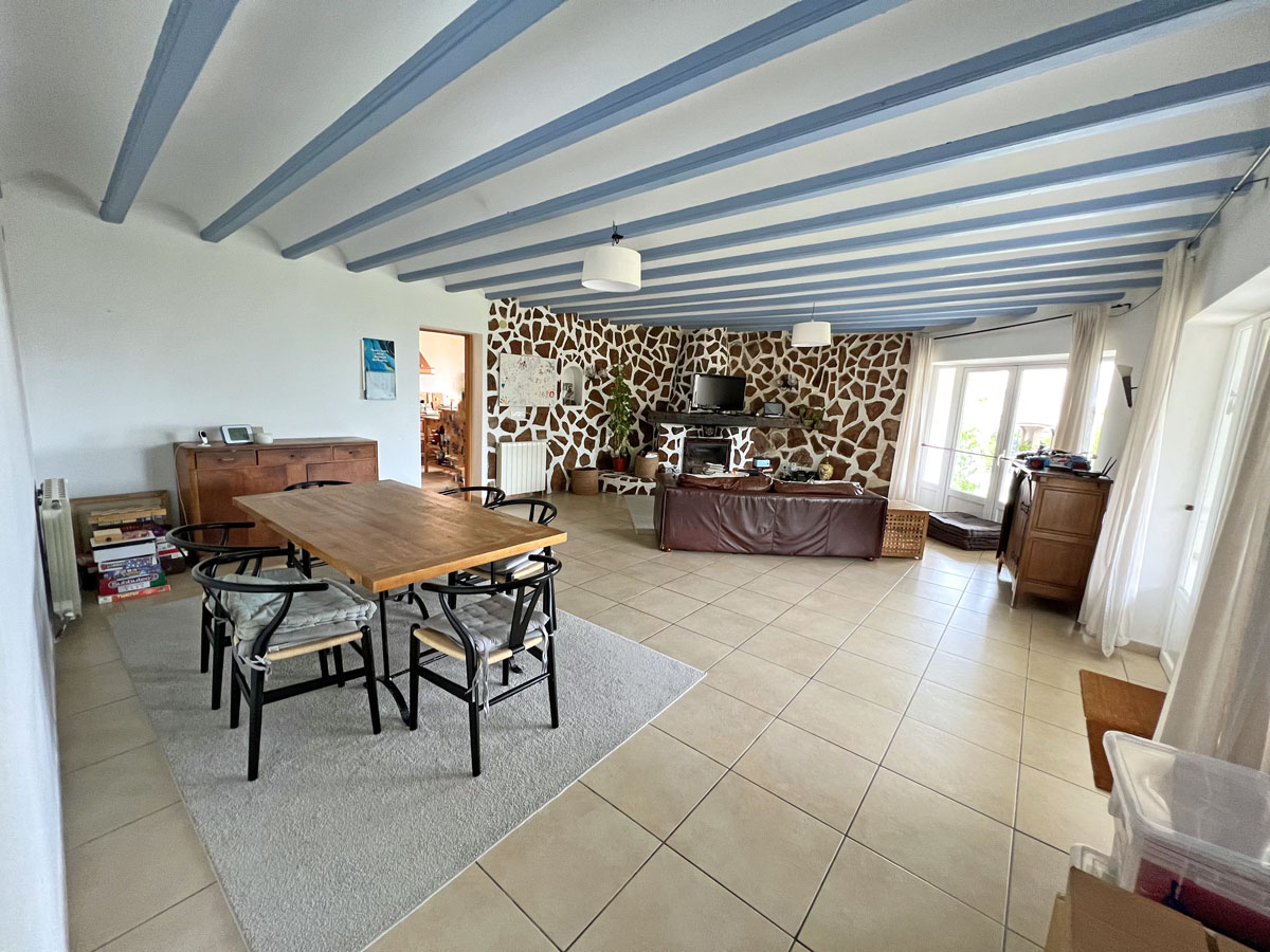 Espace de vie ouvert comprenant un salon et une salle à manger avec poutres apparentes bleues, un mur en pierre naturelle et une cheminée intégrée.