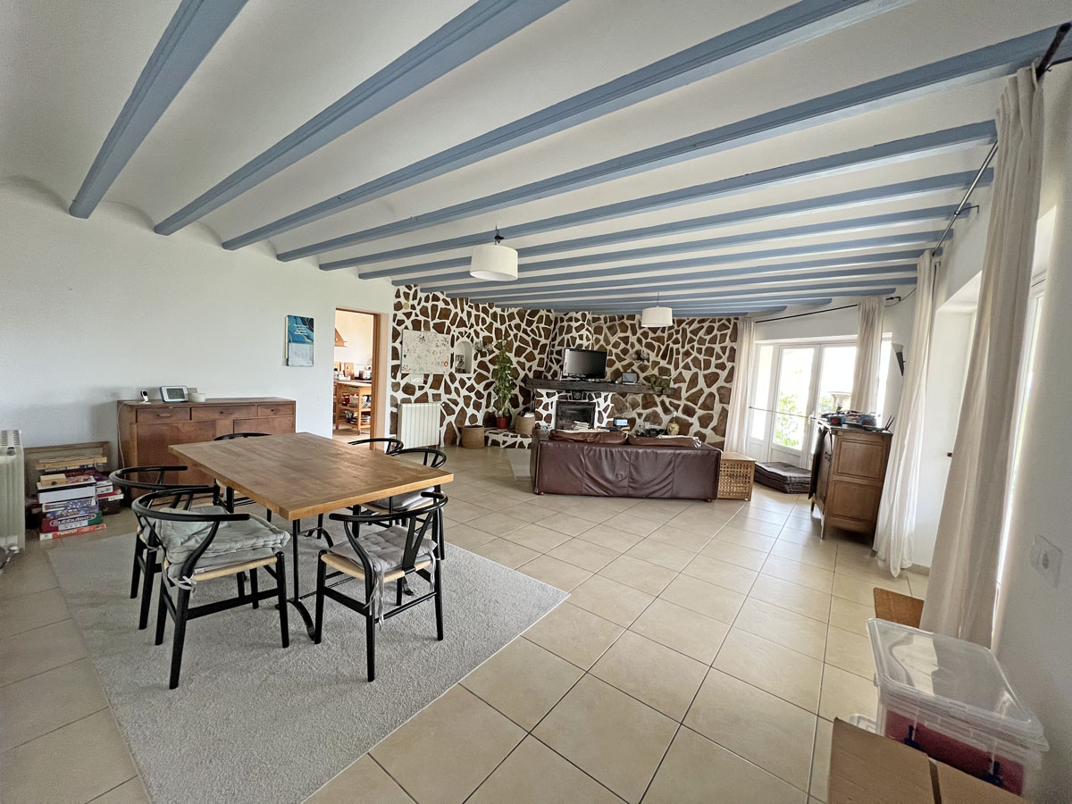 Espace de vie ouvert comprenant une salle à manger et un salon avec poutres apparentes bleu clair, un mur en pierre naturelle et un sol en carrelage beige.