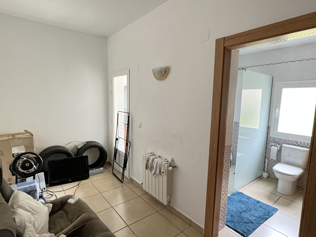 Pièce fonctionnelle avec carrelage et radiateur, attenante à une salle de bain équipée d'une douche avec paroi en verre et d'une fenêtre.